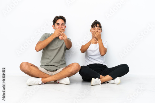 Young mixed race couple sitting on the floor isolated on white background pointing with finger at someone and laughing