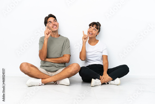 Young mixed race couple sitting on the floor isolated on white background pointing a great idea and looking up