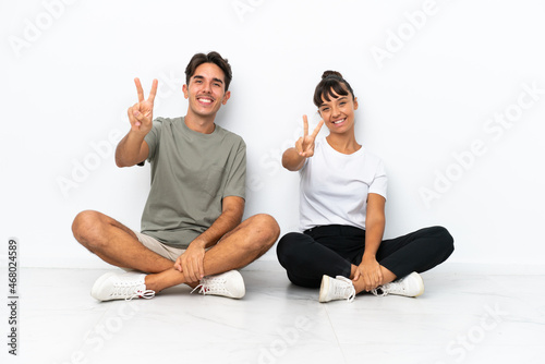 Young mixed race couple sitting on the floor isolated on white background smiling and showing victory sign