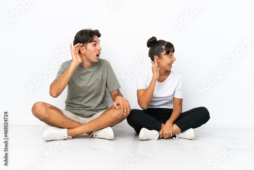 Young mixed race couple sitting on the floor isolated on white background listening to something by putting hand on the ear