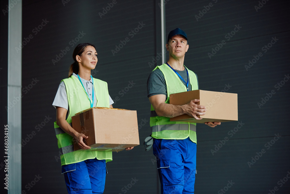 Two workers carry cardboard boxes while working at distribution warehouse. - obrazy, fototapety, plakaty 