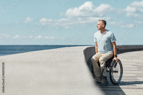 Full length portrait of adult man in wheelchair looking at water while enjoying outdoors in sunlight, copy space