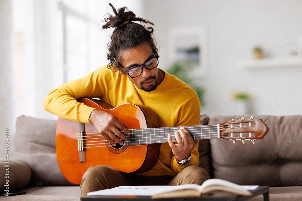 Young joyful african american man playing acoustic guitar at home, sitting on sofa in living room - obrazy, fototapety, plakaty 