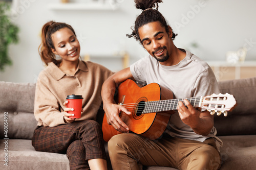Young smiling african american man musician playing instumental music on guitar for happy girlfriend