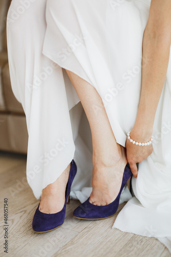 Bride in a white dress with a pearl bracelet on her arm wears blue high-heeled shoes on her feet. Close-up