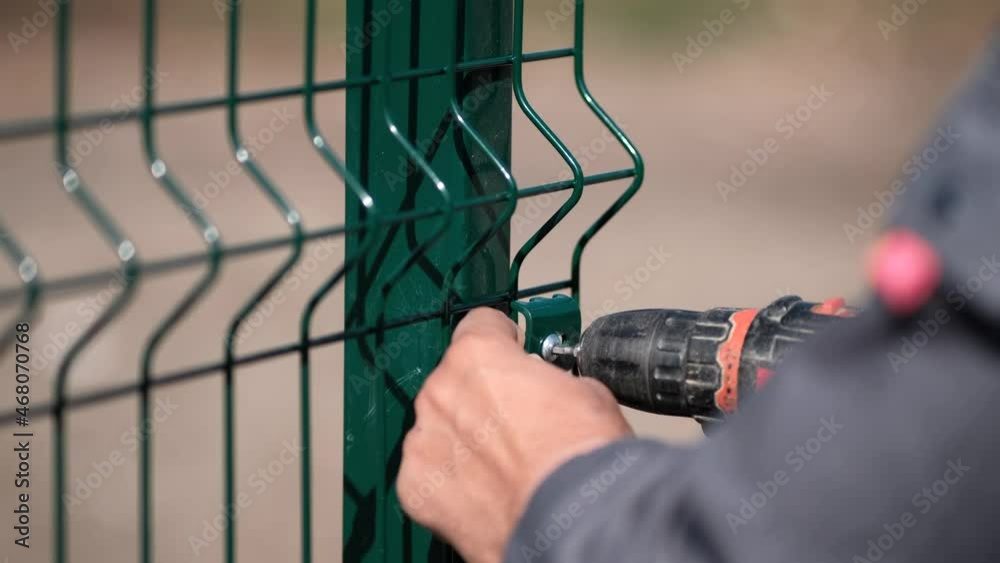 A worker screws a green mesh to a metal fence with a screwdriver. Installation of a fence in close-up.