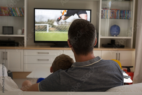 Rear view of father and son sitting at home together watching football match on tv