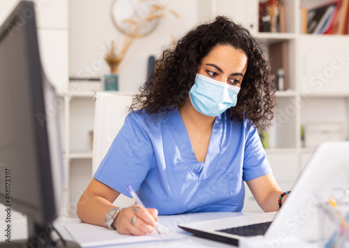Female therapist in protective mask working at table with laptop in her office