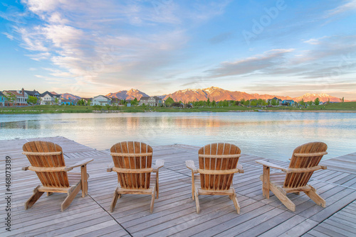 Four wooden lounge chairs facing the reflective Oquirrh Lake at Daybreak, Utah © Jason