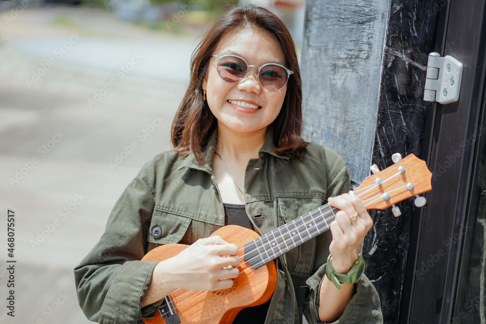 Fototapeta premium Playing Ukulele of Young Beautiful Asian Woman Wearing Jacket And Black Jeans Posing Outdoors