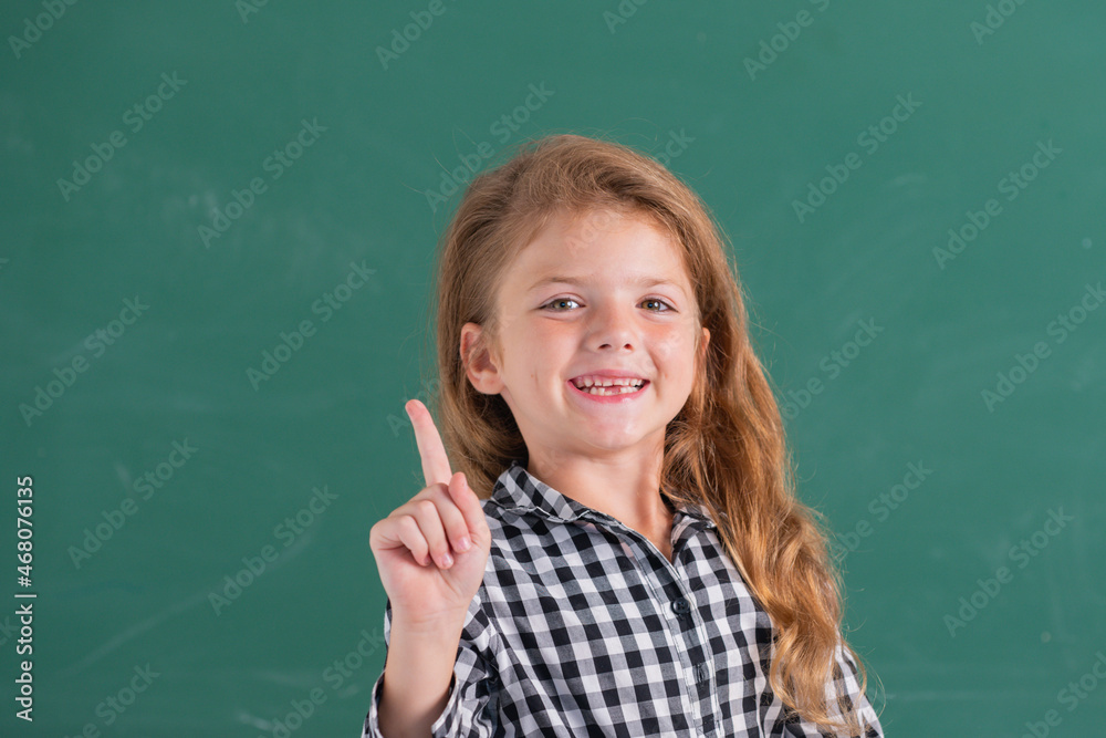 Portrait of school girl nerd pupil with surprising expression pointing with finger against blackboard.