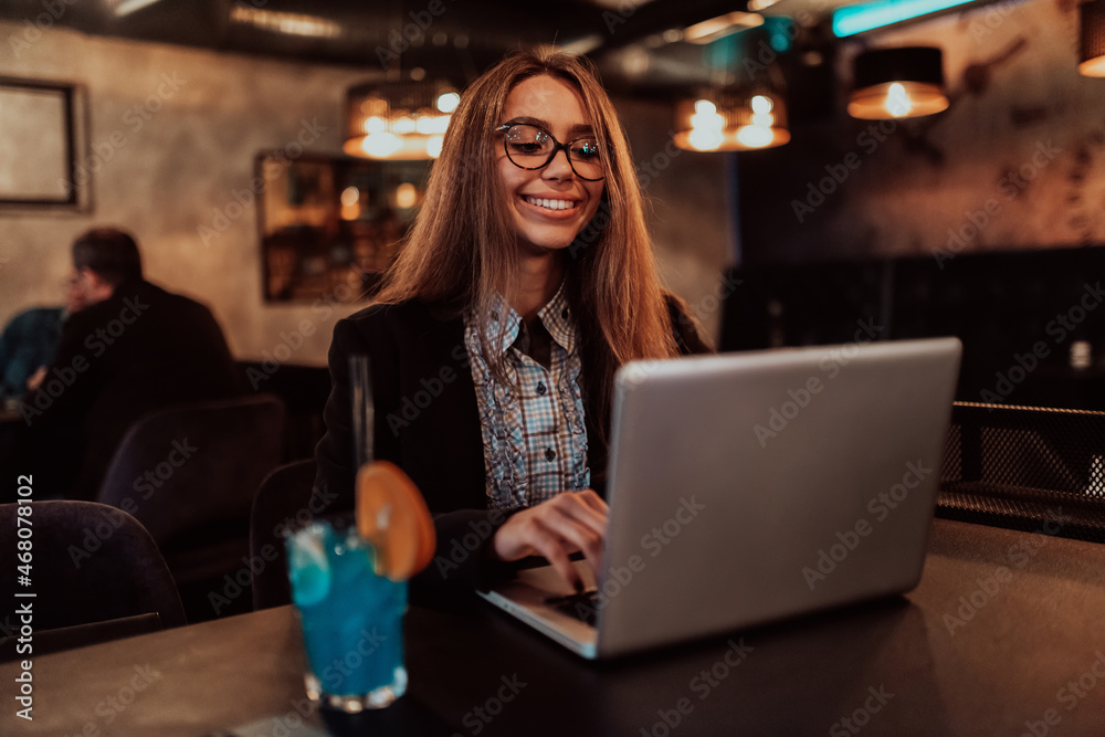 Fototapeta premium Business woman in glasses sitting in a modern cafe and working on her laptop while drinking a cocktail. Selective focus