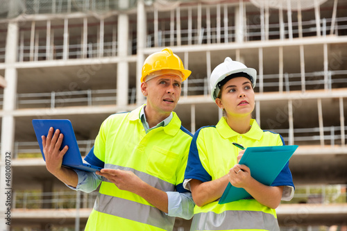 Two builders in uniform planning their work in construction plant. Man using laptop, woman holding paper folder in hands.