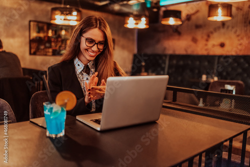 Business woman in glasses sitting in a modern cafe and working on her laptop while drinking a cocktail. Selective focus