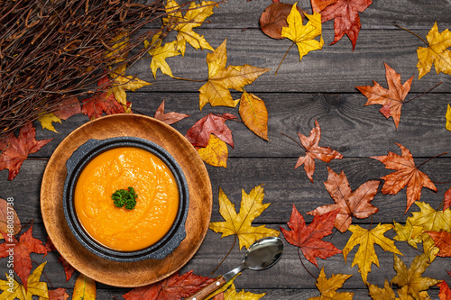 Foto Pumpkin soup in black soup tureen aside a spoon on a dark wood background with leaves and branches