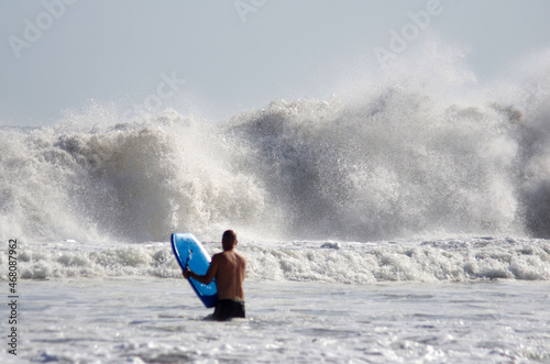 A blurred unrecognizable man's figure with a bodyboard in front of a big wave braking on a beach, vertical orientation, focus on the wave