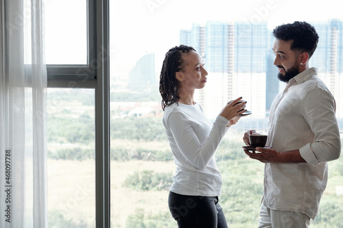 Young couple standing at big apartment window, drinking coffee and talking