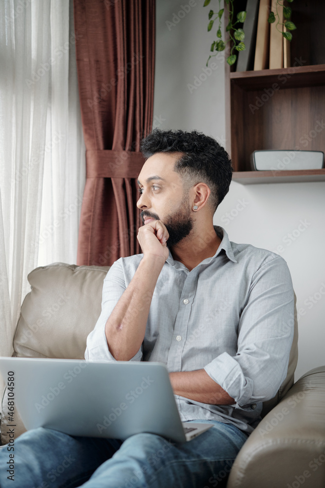 Pensive man with laptop sitting in comfy armchair and looking through window