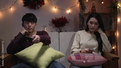 Offended asian woman sadly looking smart phone while man with remote control in hand thoughtfully face camera watching tv. Young couple in bad mood sit on sofa at modern home during valentine night
