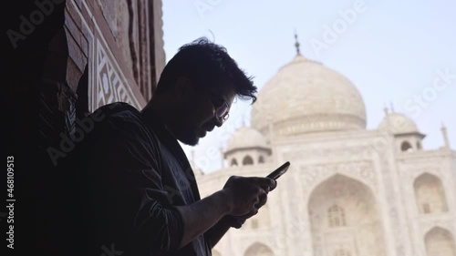 A young man or male in eye glasses sitting in front of the taj Mahal mausoleum and using a mobile phone or smartphone to type a text message. A freelance professional is browsing on a cellphone 