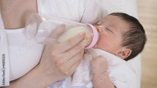 young infant is drinking milk with eyes close while its parent is using fingers to touch the bottle lightly in the bedchamber with cropped shot.