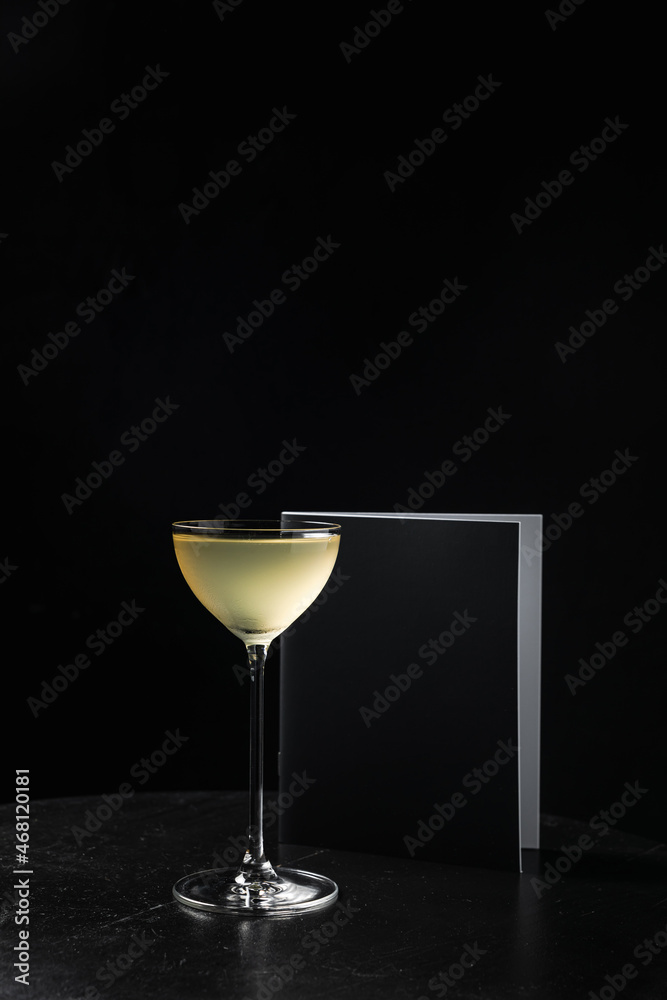 An alcoholic cocktail in a nick and nora glass and a menu book on black wooden table, black background