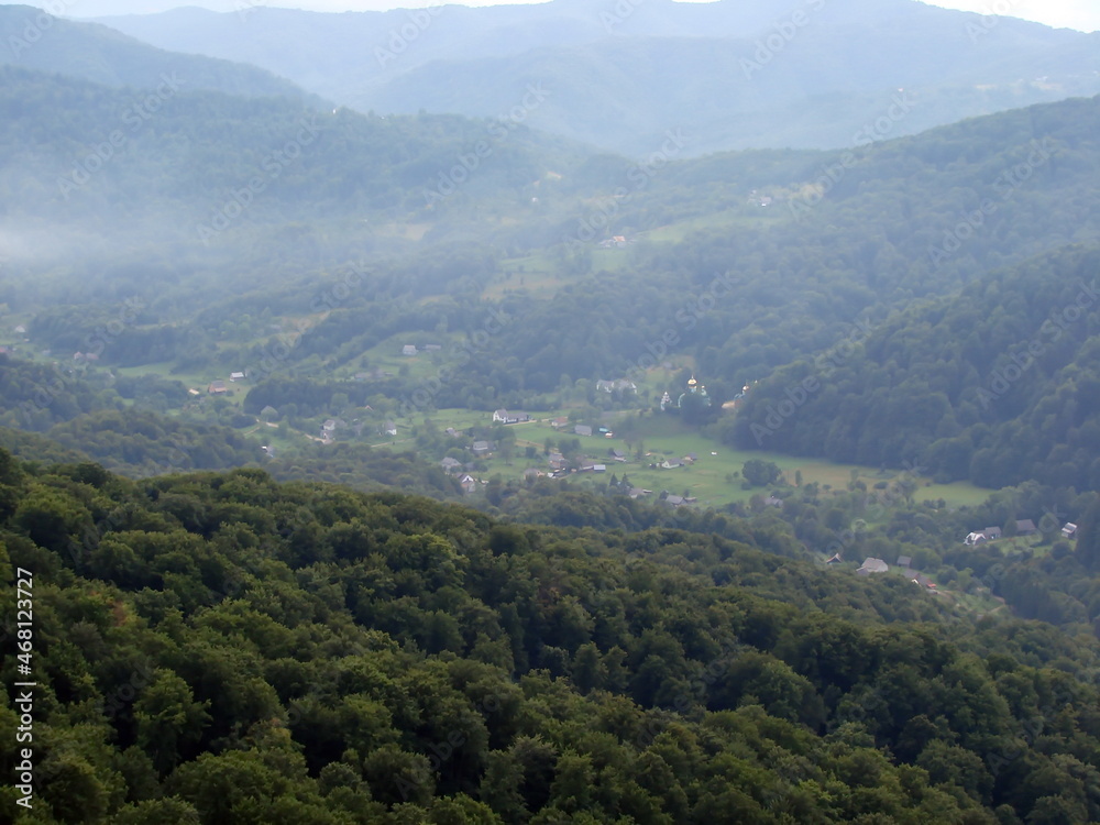 Landscape of a mountain village surrounded by green waves of beech forests under the fog hanging in the air.
