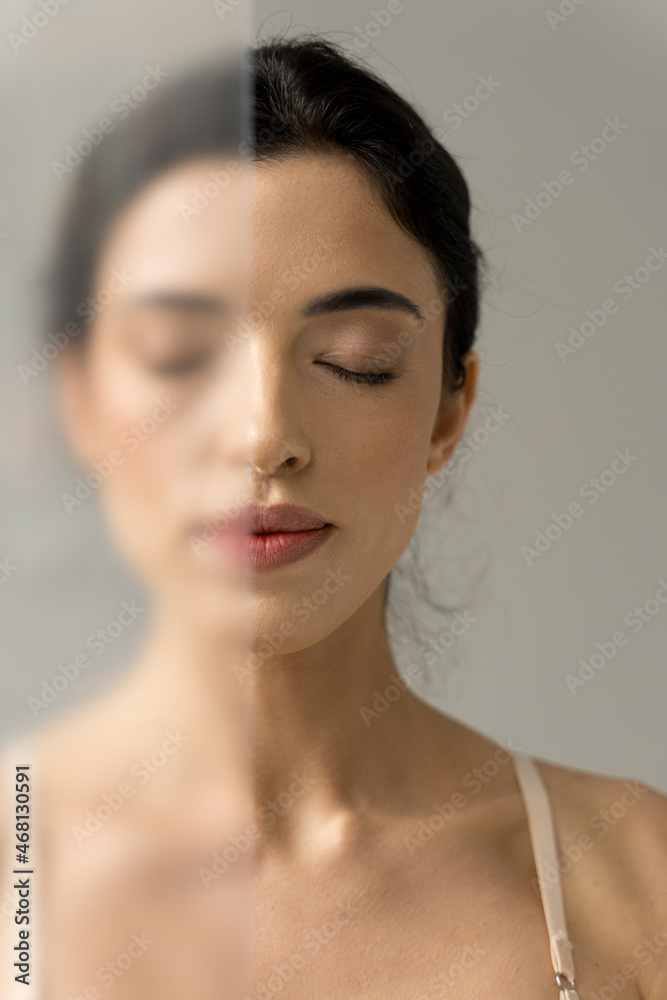 Naklejka premium Woman standing with closed eyes behind the transparent glass over the white studio wall