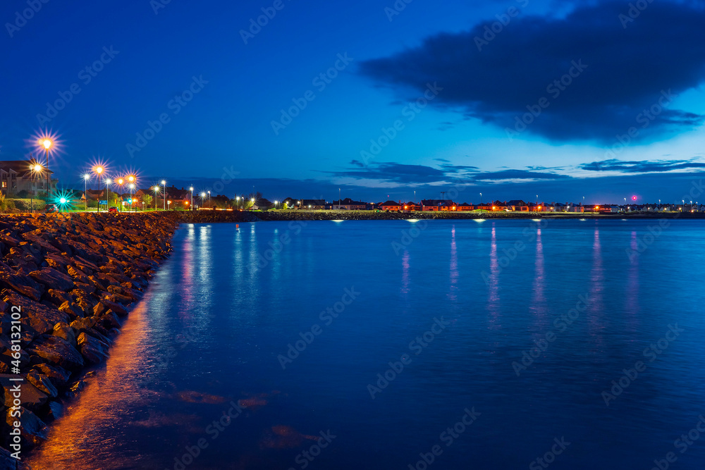 Salthill promenade waterfront before sun rise. Galway city, Ireland. Blue and orange color. Cloudy sky. Nobody. City lights and reflection in water surface.