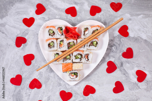 Sushi rolls with salmon on white heart-shaped plate and wooden sushi sticks among red confetti hearts
