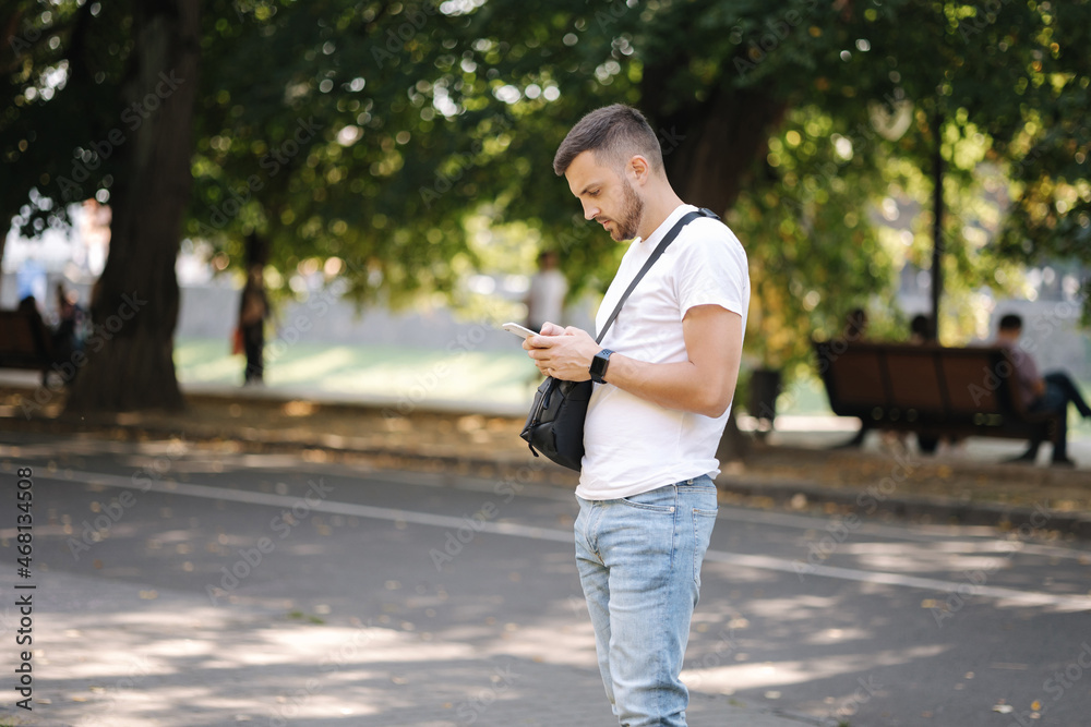 Fototapeta premium Man in white t-shirt with black shouler bag using phone outddor. Man has smart watch