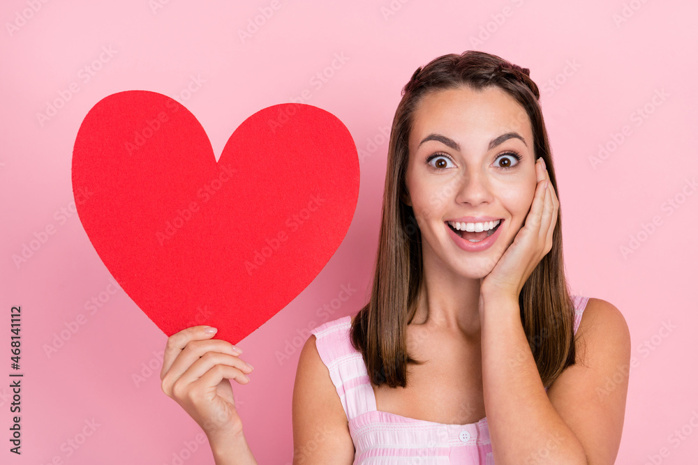 Photo of amazed happy young woman hold red paper heart finger hand cheek isolated on pink color background Photo of amazed happy young woman hold red paper heart finger hand cheek isolated on pink color background