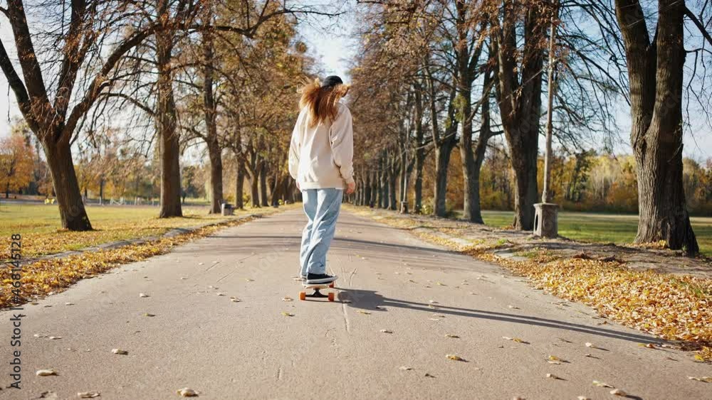 Female skateboarder beginer learns to ride a long skate in a city park. Skateboarding training. Back view, autumn sunny day