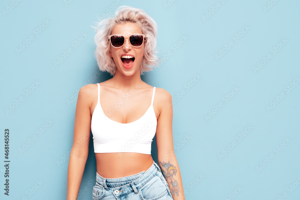 Young beautiful smiling female in trendy summer clothes. Sexy carefree woman posing near blue wall in studio. Positive blond model having fun in sunglasses. Screaming and shouting Young beautiful smiling female in trendy summer clothes. Sexy carefree woman posing near blue wall in studio. Positive blond model having fun in sunglasses. Screaming and shouting