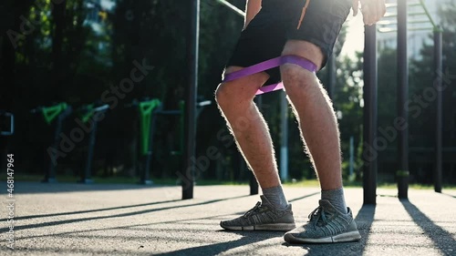 Man guy doing squats with elastic rubber band at the stadium outdoors. Athlete male person during workout with additional sport equipmant photo