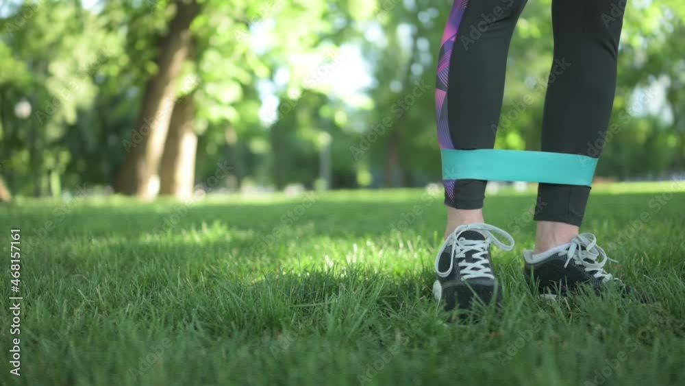 Girl with elastic rubber band training her legs in park outdoors. Young woman exercising at nature with sport equipment closeup view Girl with elastic rubber band training her legs in park outdoors. Young woman exercising at nature with sport equipment closeup view