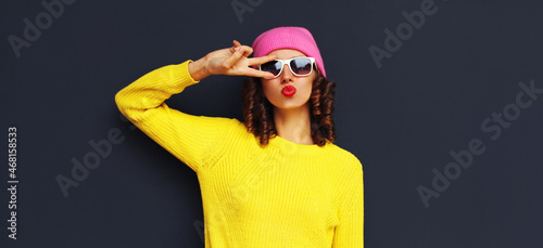 Portrait of stylish young woman blowing her lips sending sweet air kiss wearing an yellow knitted sweater and pink hat on dark background