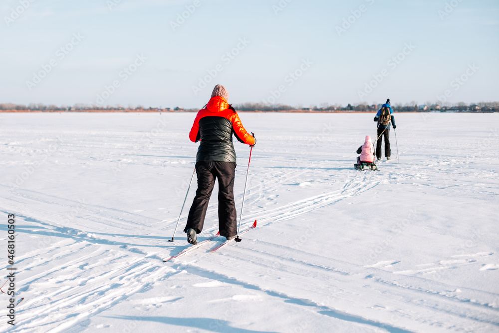 Family on ski trip. Young woman, man and child are skiing in winter on frozen river near forest, man is sledding child. 