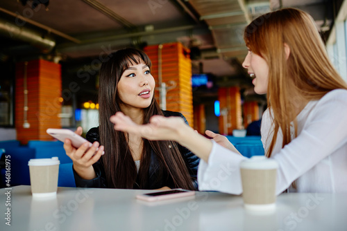 Asian and Caucasian female bloggers discussing content text during leisure in coffee shop, diverse millennial women talking about social media networking have friendly meeting in cafe interior