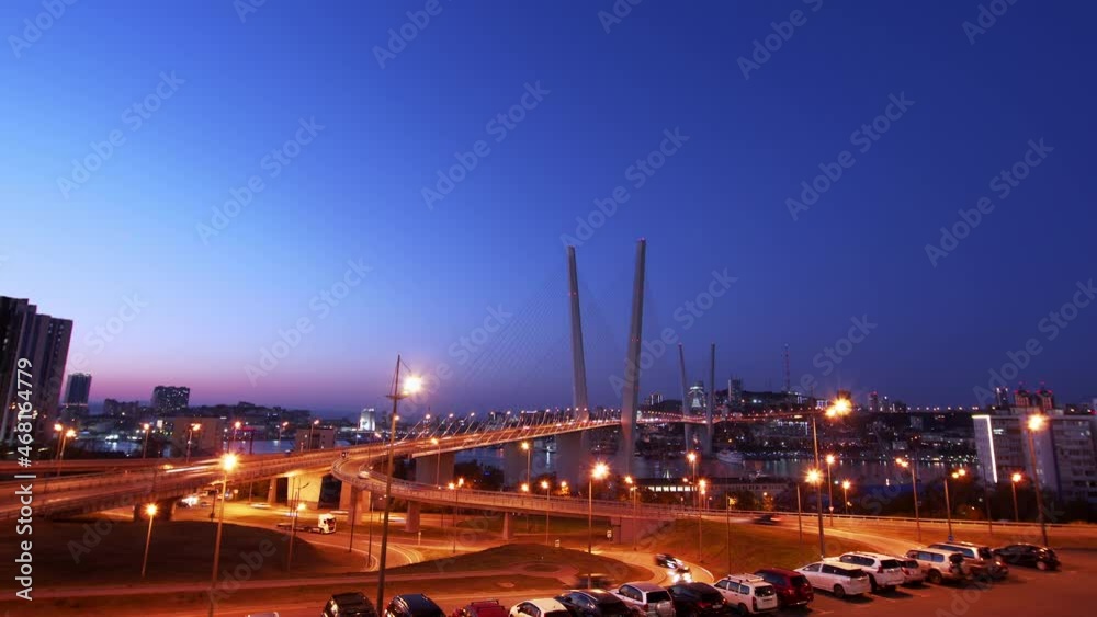 Timelapse of sunset above the illuminated Golden bridge across the Zolotoy Rog (Golden Horn Bay) in Vladivostok, Russia. Cityscape, busy roads, car parking. The sky is getting dark. Evening. 