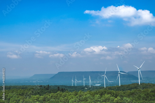 The big white wind turbine in farm for spinning electricity sees large mountains far away, with clouds and blue skies above the mountains.