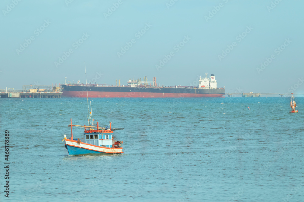 A small blue fishing boat was sailing in the middle of the sea, heading for the shore. There is a sky and a large cargo ship as a backdrop.