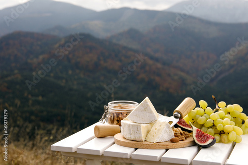 Delicious cheese, nuts and fruits on white wooden table against mountain landscape. Space for text