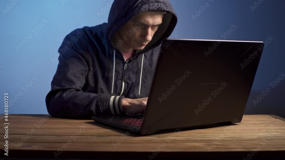 A hooded hacker is typing on a laptop keyboard in a dark room under a blue light. The concept of cybercrime fraud and identity theft