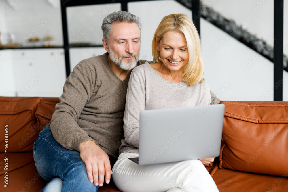 Middle-aged couple spends leisure time with a laptop at home. Spouses sit on the couch, watching video, web surfing or shopping online together, enjoying time online Middle-aged couple spends leisure time with a laptop at home. Spouses sit on the couch, watching video, web surfing or shopping online together, enjoying time online