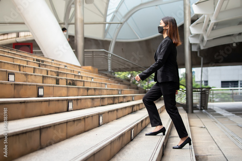 A long-haired woman wearing a mask, suit jacket, trousers and black shoes is walking up the stairs.