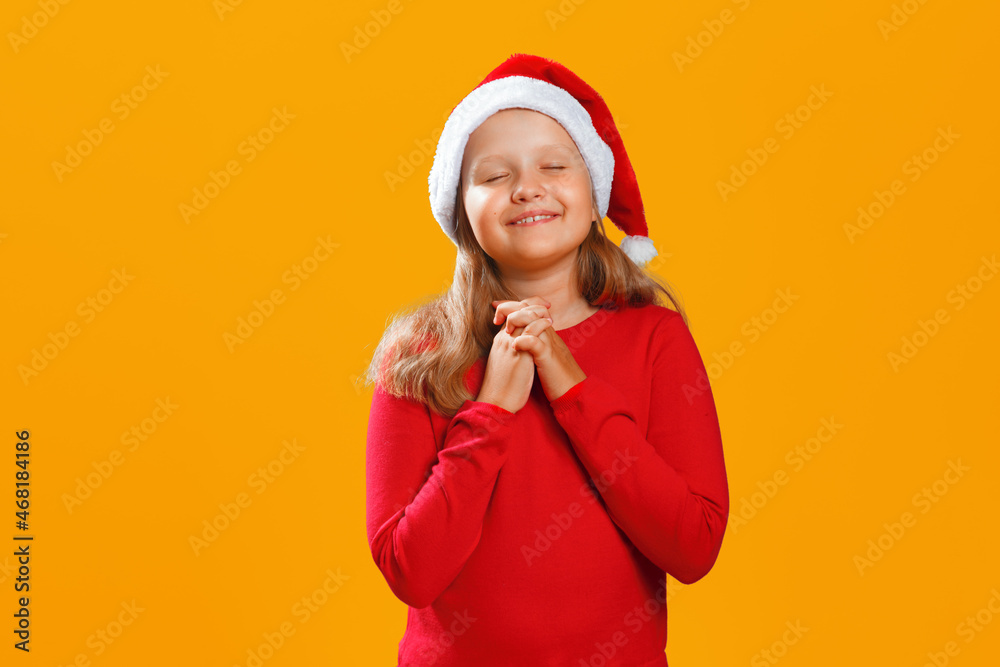 Little girl in santa hat is waiting for christmas. The child closed his eyes and made a wish on a yellow background. Little girl in santa hat is waiting for christmas. The child closed his eyes and made a wish on a yellow background.