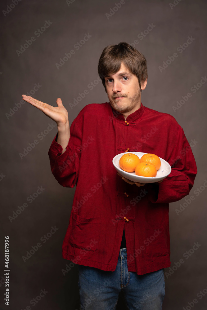 Young handsome tall slim white man with brown hair not knowing how to feel about his oranges in red shirt on grey background Young handsome tall slim white man with brown hair not knowing how to feel about his oranges in red shirt on grey background