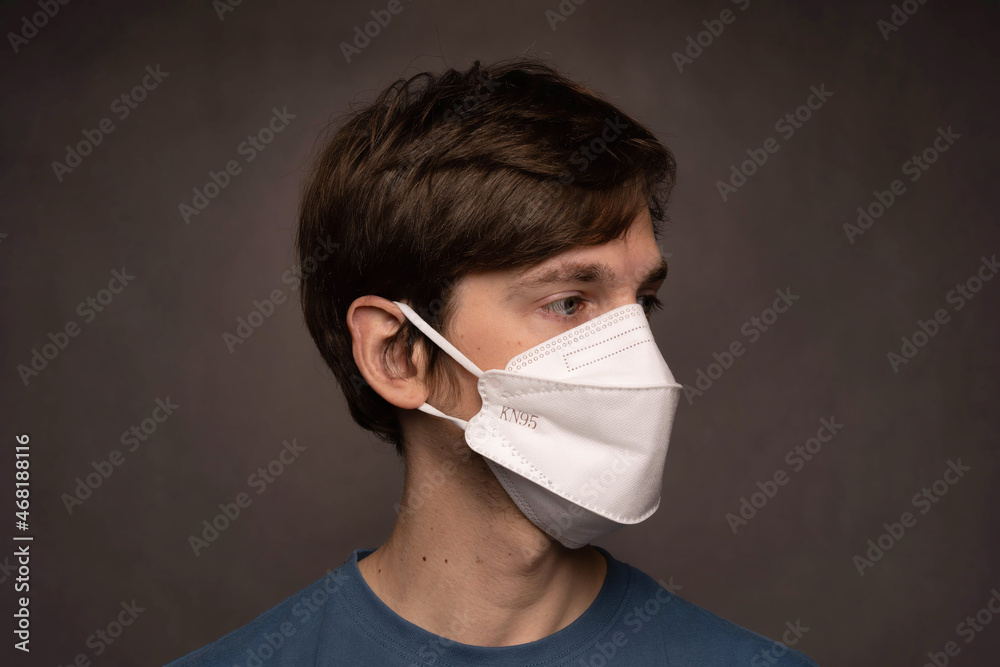 Young handsome tall slim white man with brown hair looking right with face mask on in blue shirt on grey background Young handsome tall slim white man with brown hair looking right with face mask on in blue shirt on grey background