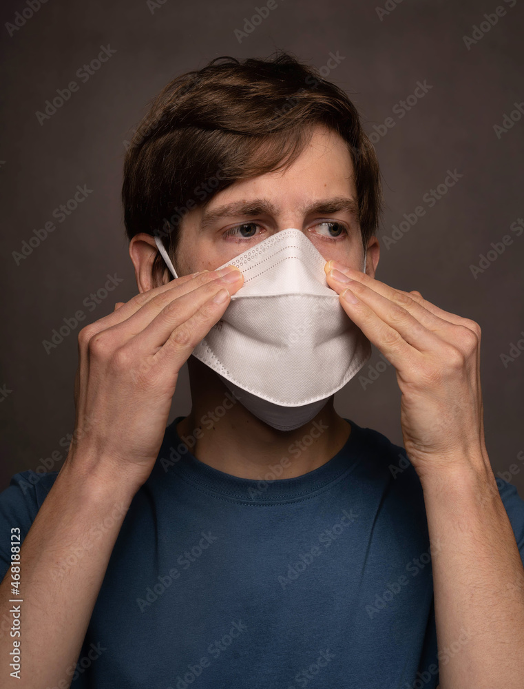 Young handsome tall slim white man with brown hair with both hand on face mask in blue shirt on grey background Young handsome tall slim white man with brown hair with both hand on face mask in blue shirt on grey background